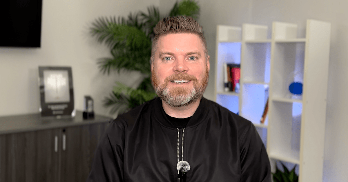 Andrew Aebersold, CEO of Mediaura, looks into the camera while explaining how cutting out marketing during a downturn can hurt your business. He is wearing a black zip up. The background is an office scene, featuring a bookshelf and plant.