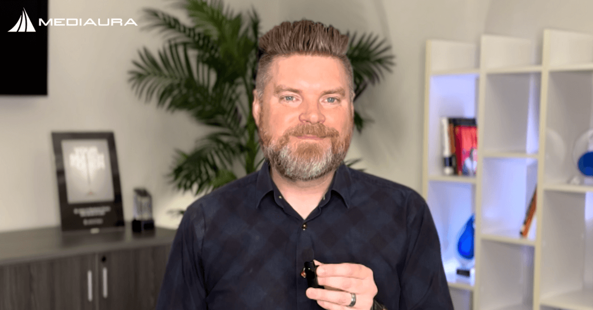 Andrew Aebersold, CEO of Mediaura, looks into the camera while explaining ROI vs ROAS. He is wearing a dark blue checkerboard print collared shirt. The background is an office scene, featuring a bookshelf and plant.