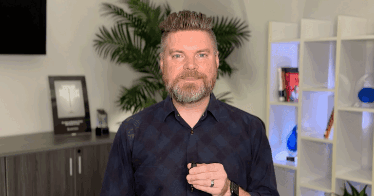 Andrew Aebersold, CEO of Mediaura, looks into the camera while explaining what to look for when hiring a digital marketer. He is wearing a dark blue checkerboard print collared shirt. The background is an office scene, featuring a bookshelf and plant.