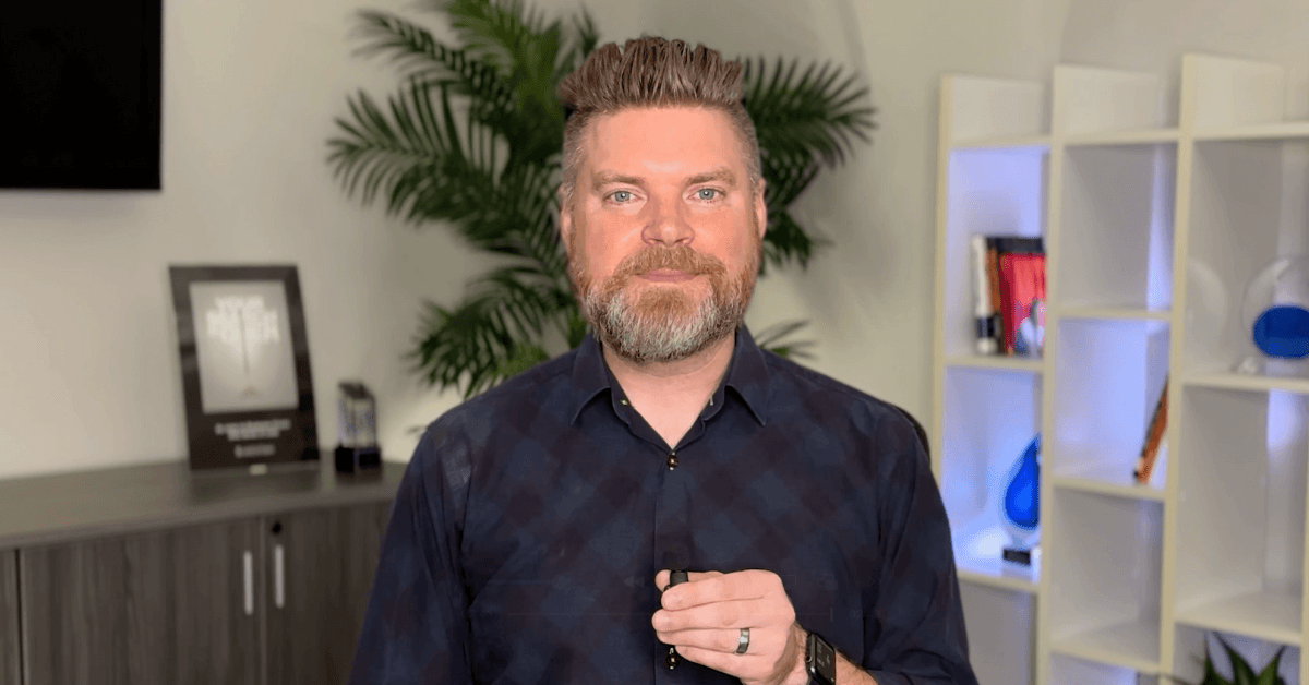 Andrew Aebersold, CEO of Mediaura, looks into the camera while explaining what to look for when hiring a digital marketer. He is wearing a dark blue checkerboard print collared shirt. The background is an office scene, featuring a bookshelf and plant.