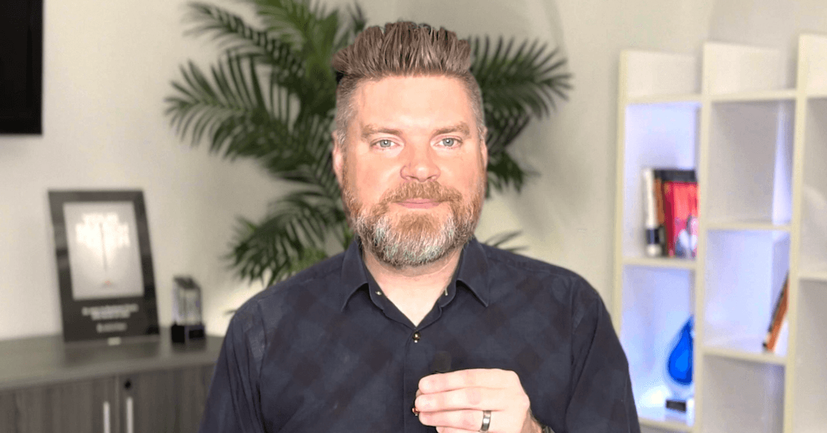 Andrew Aebersold, CEO of Mediaura, looks into the camera while explaining why your AI marketing questions may be getting bad results. He is wearing a dark blue checkerboard print collared shirt. The background is an office scene, featuring a bookshelf and plant.