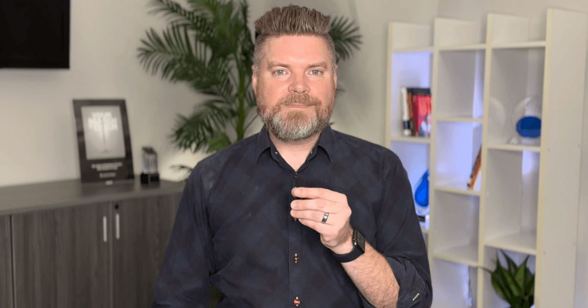 Andrew Aebersold, CEO of Mediaura, looks into the camera while explaining why you need your marketing funnel in place before running awareness ads. He is wearing a dark blue checkerboard print collared shirt. The background is an office scene, featuring a bookshelf and plant.