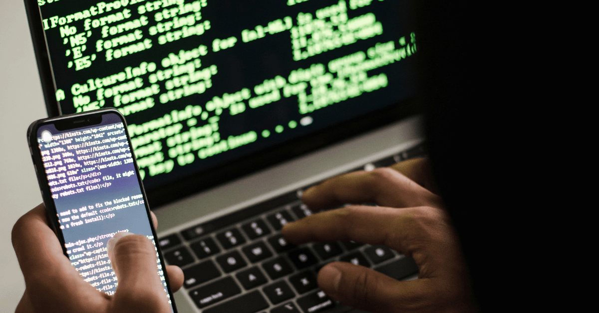 An anonymous hooded hacker figure holds a smart phone and types on a laptop, both featuring lines of code. A website hack puts your marketing at risk, damaging your reputation and tanking SEO and online ad campaigns.