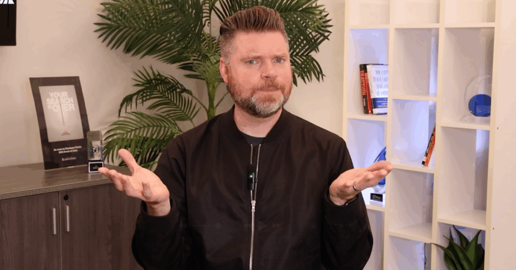 Andrew Aebersold, CEO of Mediaura, looks into the camera while explaining who to credit for your leads: sales or marketing. He is wearing a black zip-up. The background is an office scene, featuring a bookshelf and plant.