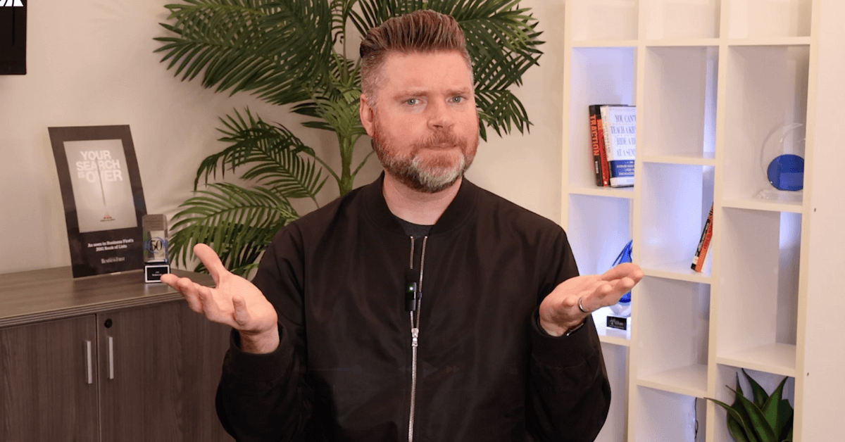 Andrew Aebersold, CEO of Mediaura, looks into the camera while explaining who to credit for your leads: sales or marketing. He is wearing a black zip-up. The background is an office scene, featuring a bookshelf and plant.