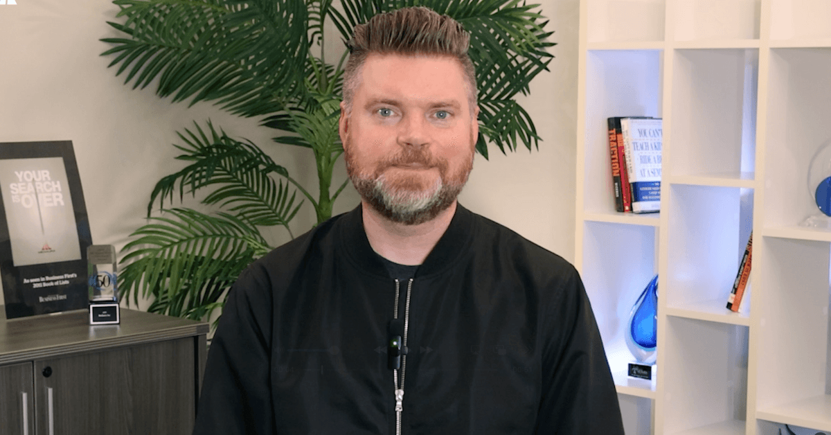 Andrew Aebersold, CEO of Mediaura, looks into the camera while explaining how behavioral healthcare seasonality is totally normal and how to get through it. He is wearing a black zip-up. The background is an office scene, featuring a bookshelf and plant.