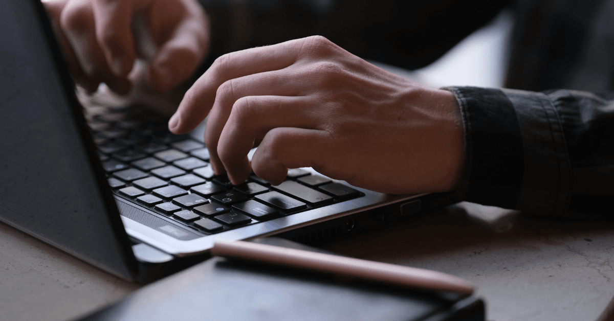 A closeup of someone typing on a black laptop, wearing a long-sleeve black button down, with a black notebook in the foreground, representing a competitor buying your name on Google.